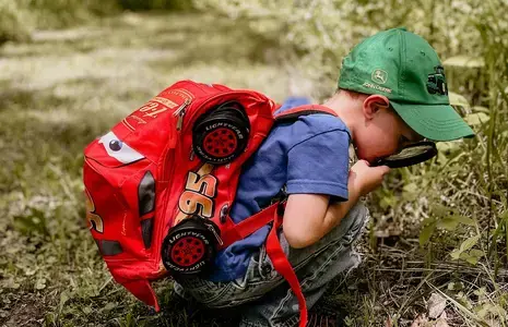 Rohelise nokamütsi ja punase seljakotiga poiss uurib luubiga loodust